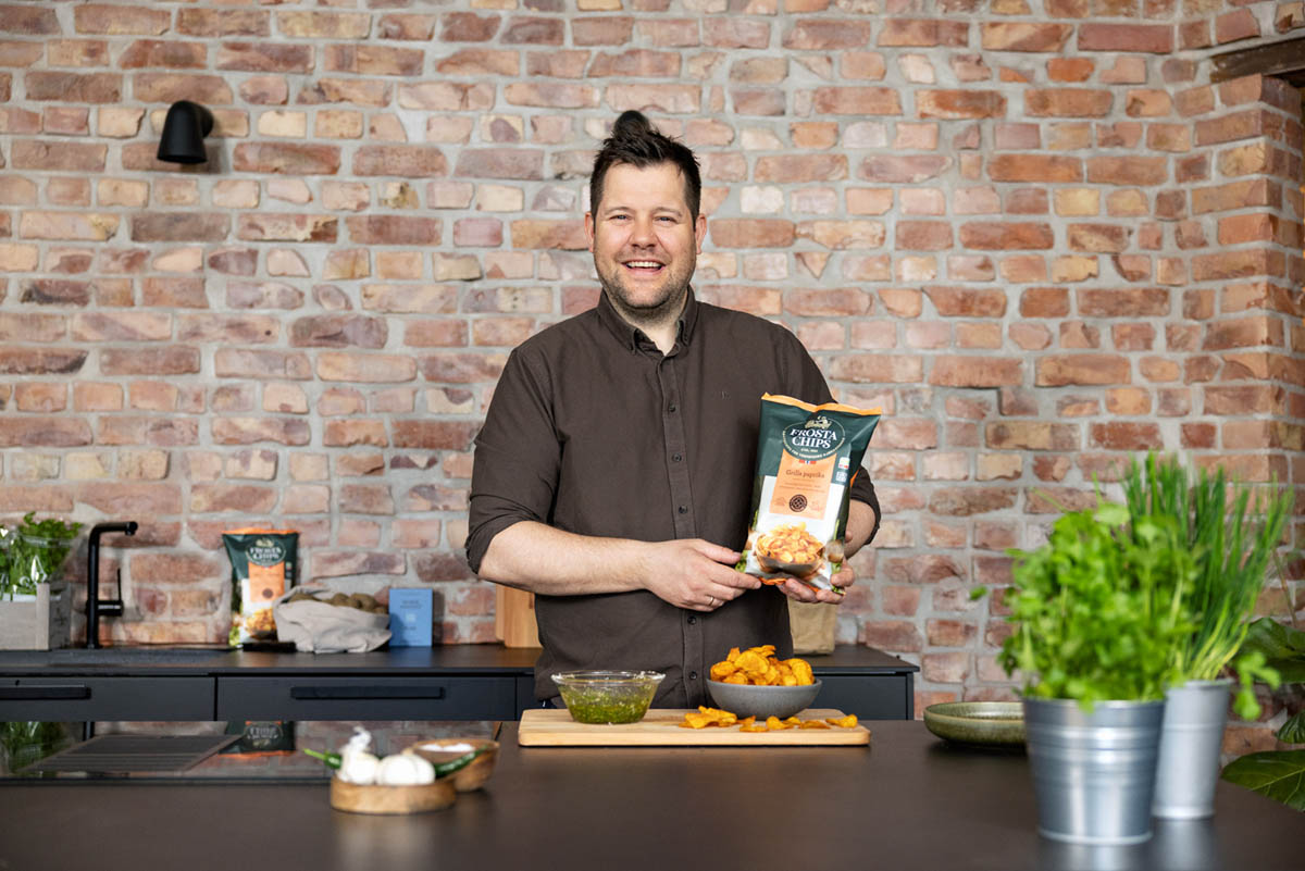 En kokk holder opp en pose chips i et rustik kjøkken, ser og smiler i kamera