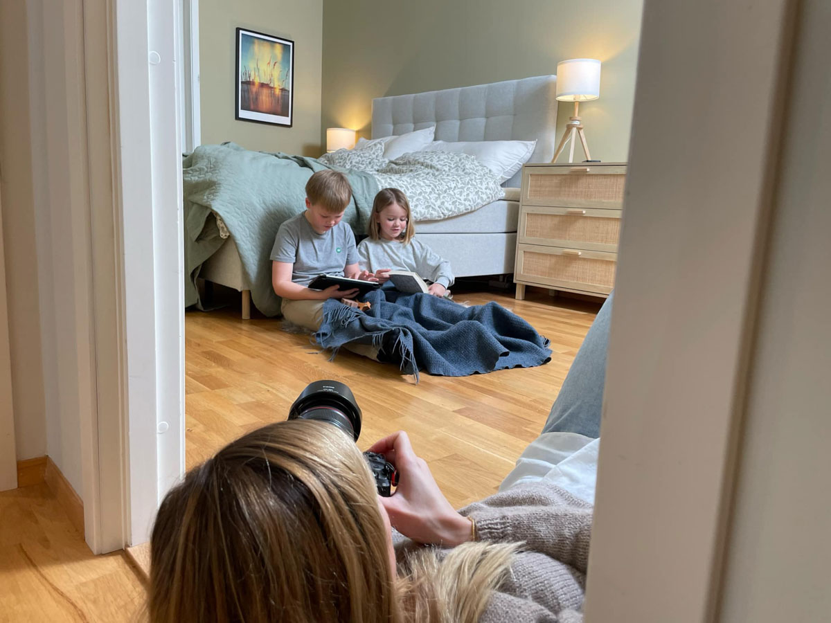 To barn leser bok og ser på ipad mens de sitter på gulvet lent mot en seng, i forgrunnen ser vi en kvinne som tar bilde av dem