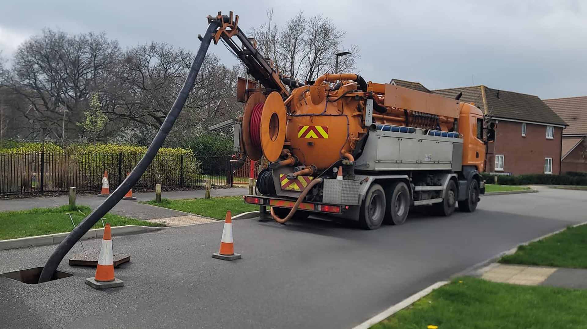 Orange vacuum truck with large hose extended into an open manhole on a residential street.