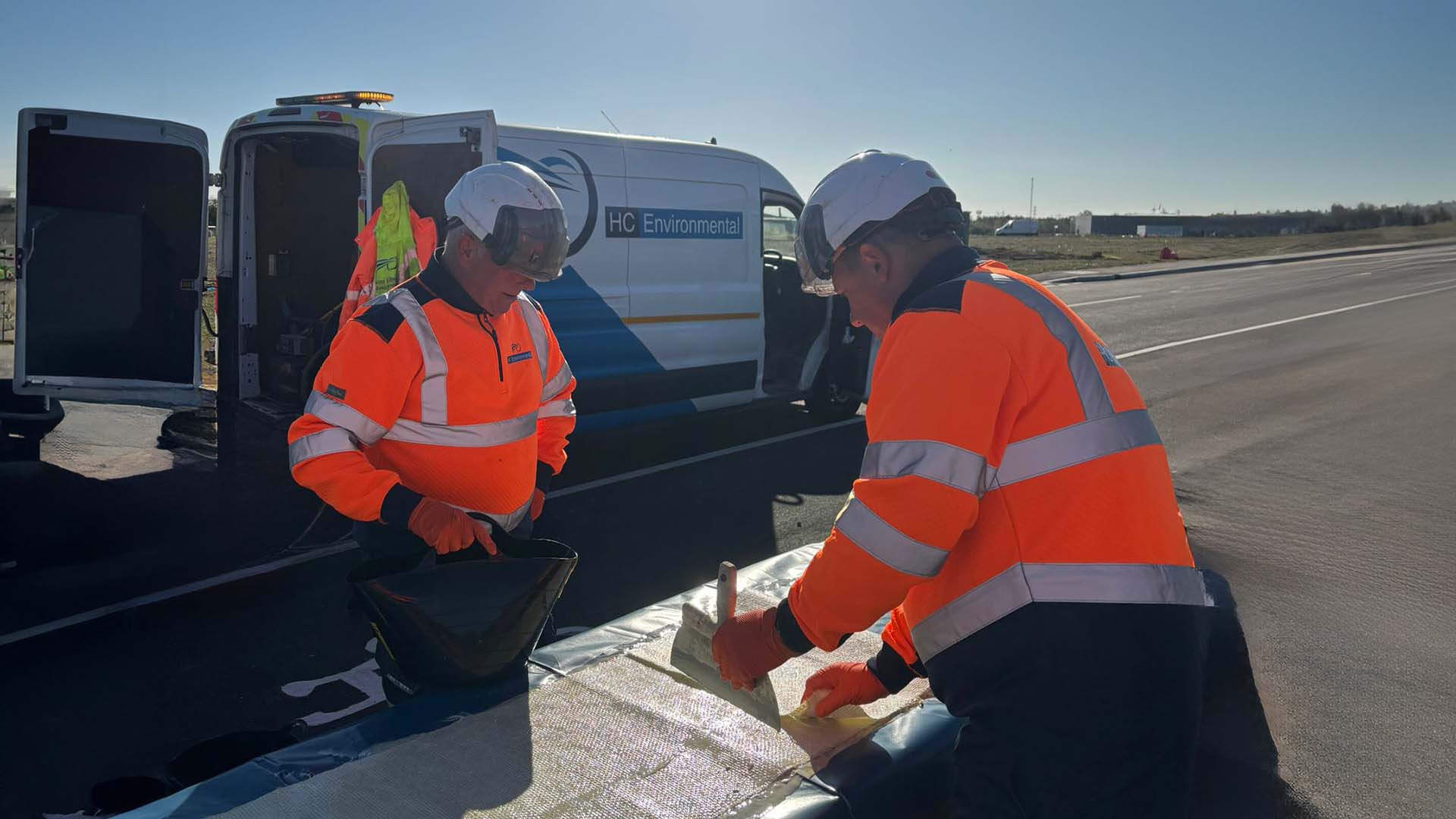 Two workers in high-visibility orange jackets and helmets applying a material on a flat surface near a white van labeled HC Environmental on a sunny day.
