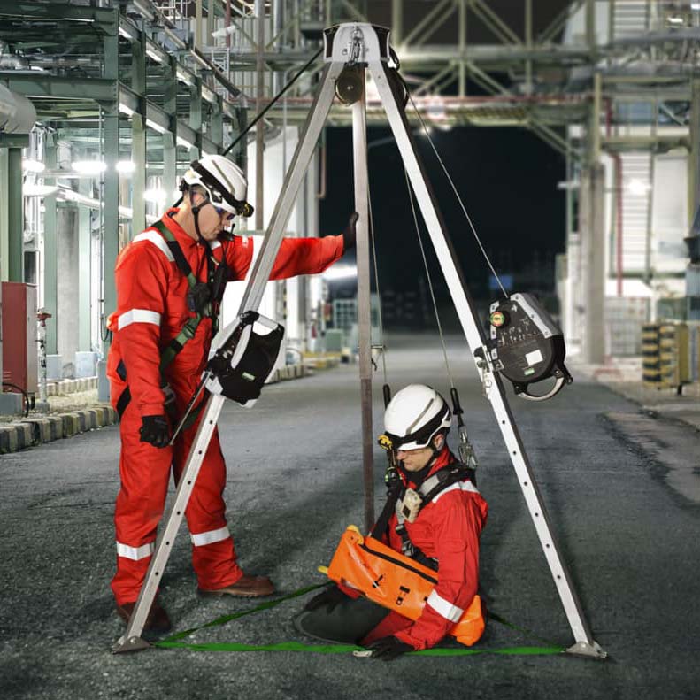 Two workers in red safety suits and helmets using a tripod and harness system for confined space entry at an industrial site at night.