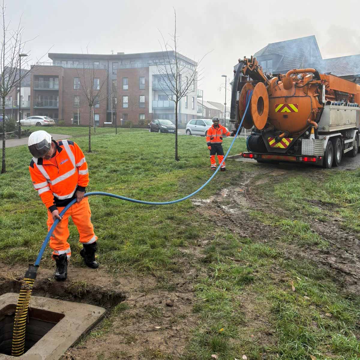 Two workers in orange high-visibility clothing operating a blue hose connected to an orange jetting truck near an open manhole in a grassy area.