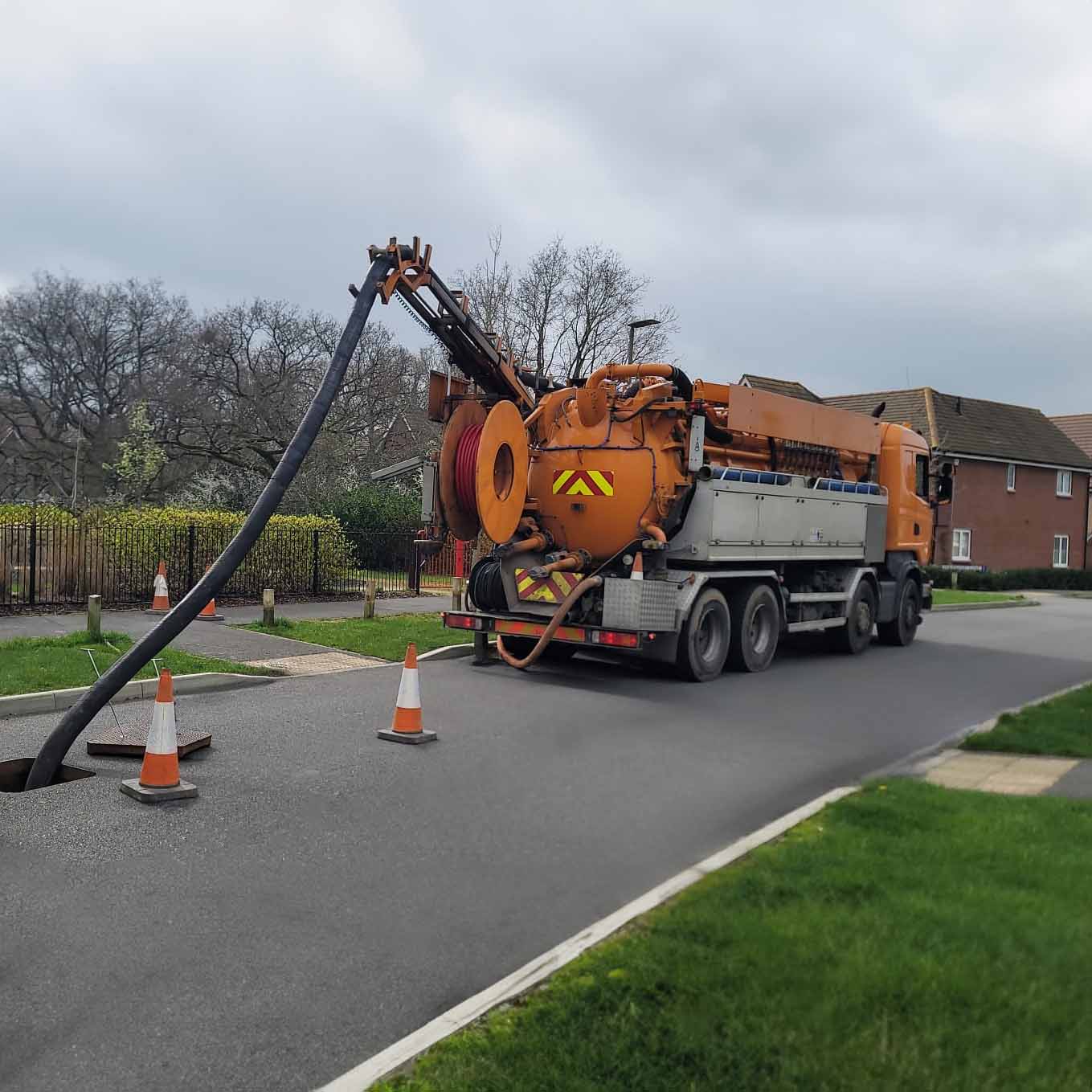 Orange vacuum excavation truck parked on a residential street with a large hose inserted into a manhole, surrounded by safety cones.