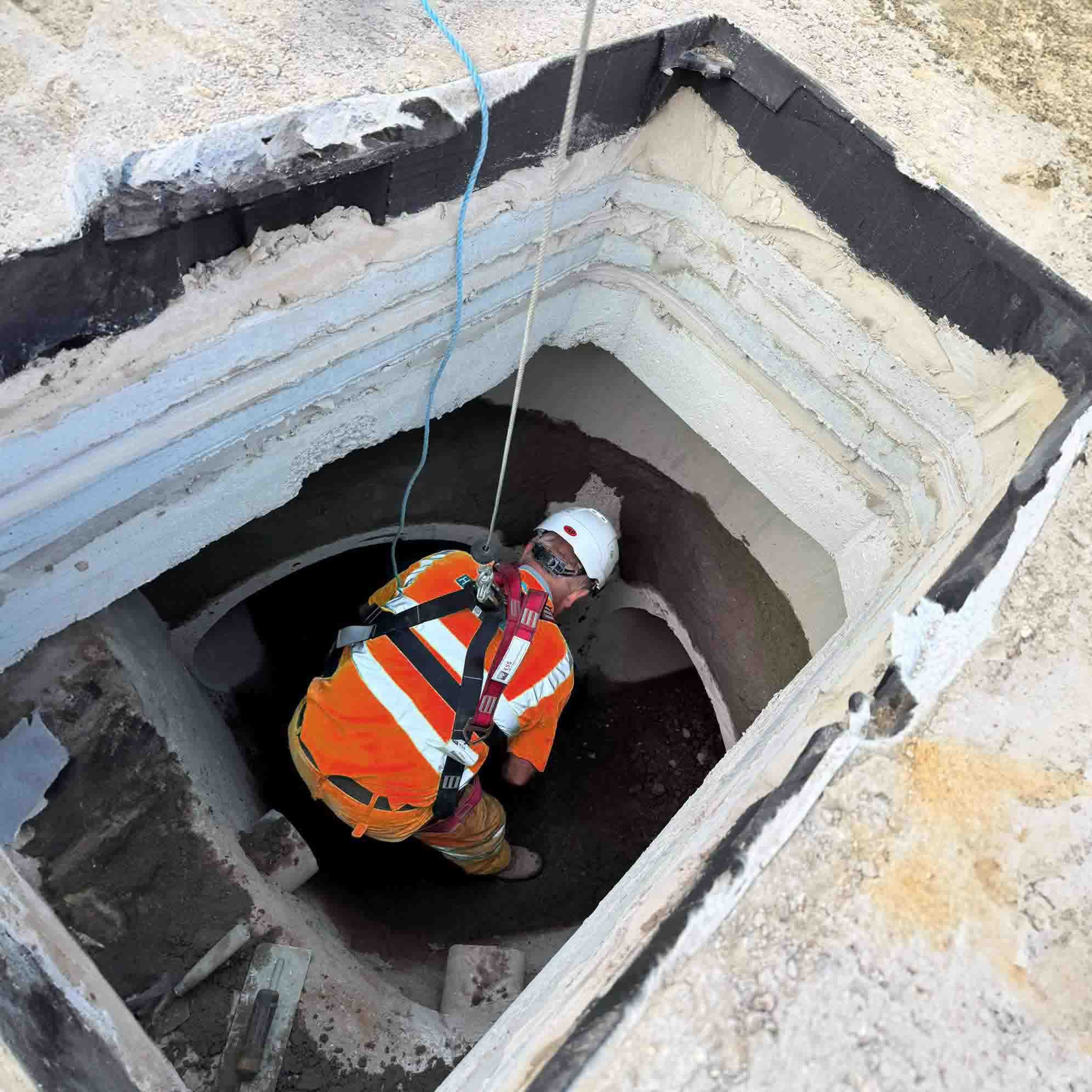 Worker in an orange safety vest and white helmet descending into a deep rectangular manhole with layered concrete walls, secured by a safety harness.