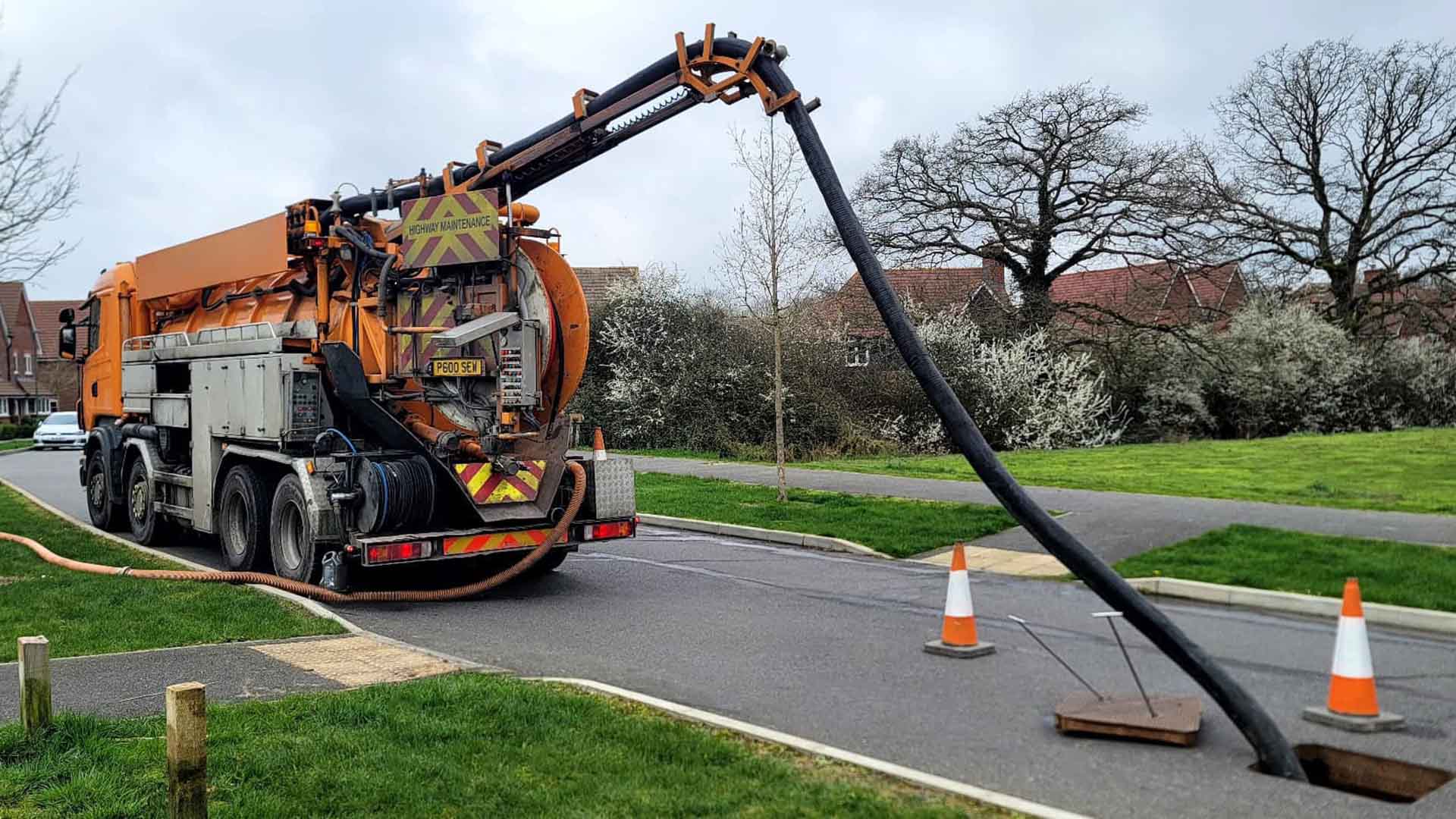Orange highway maintenance vacuum truck with large suction hose extending into a street manhole, surrounded by safety cones.