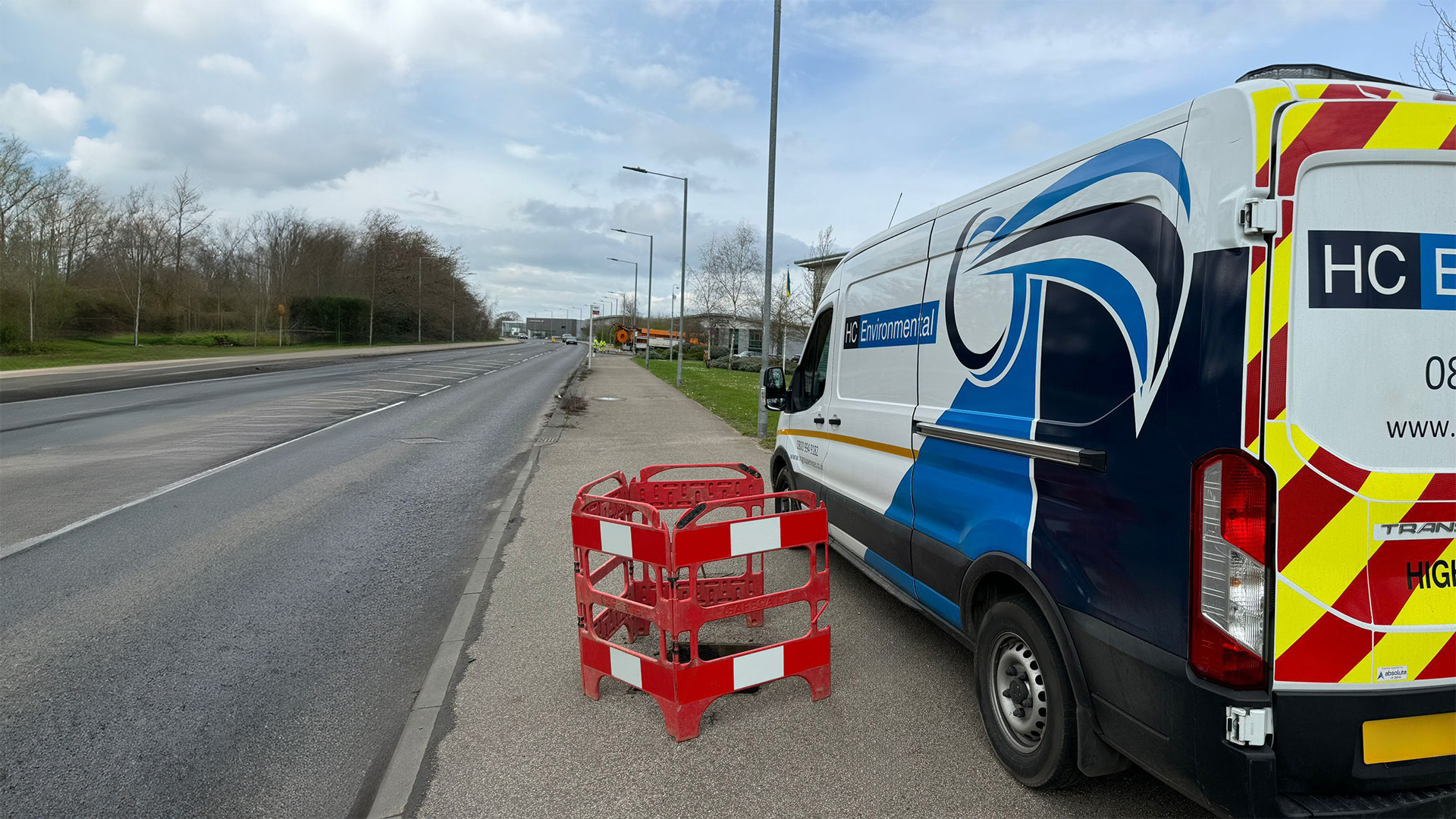White and blue HC Environmental van parked on the side of a road next to red safety barriers covering a hole in the pavement.