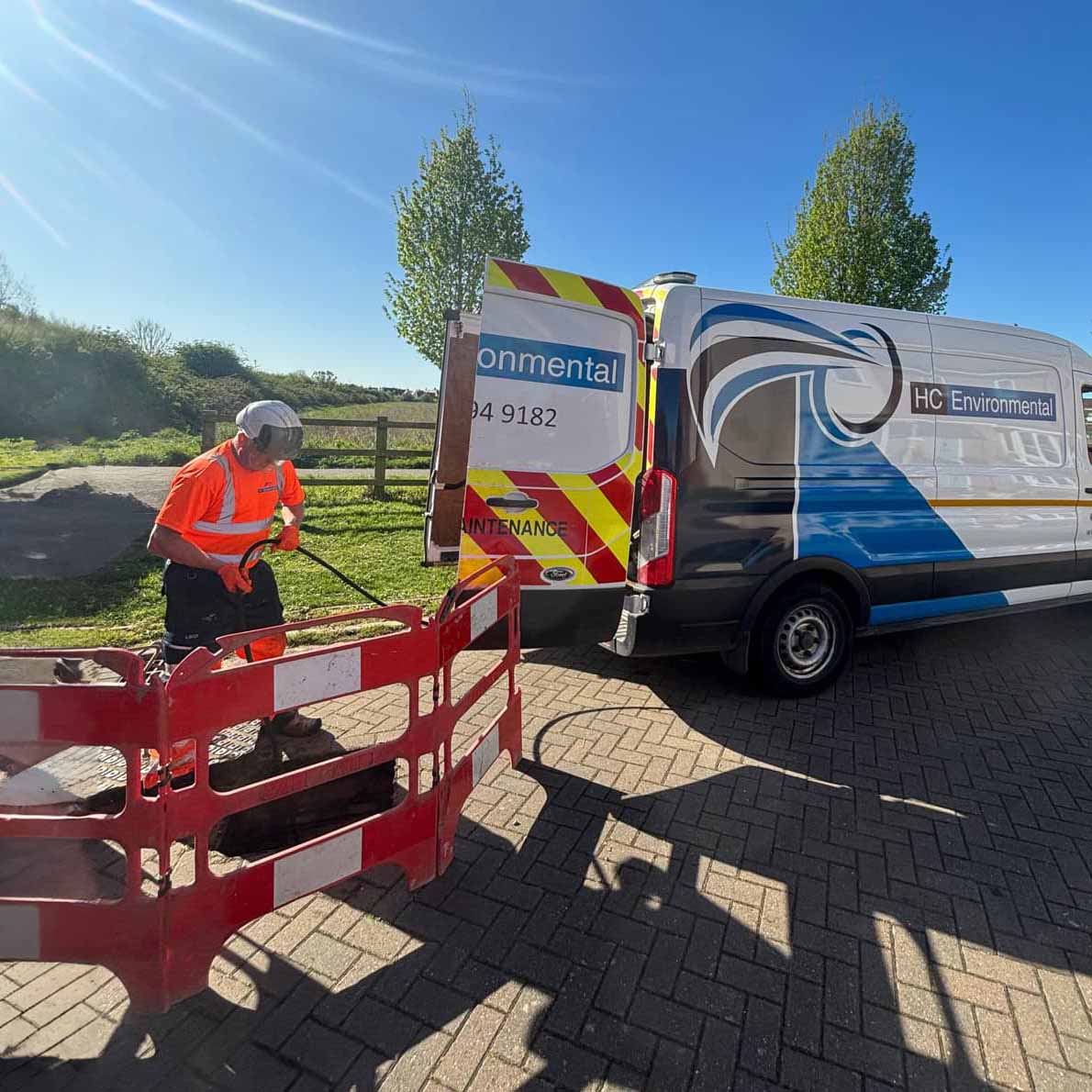 Worker in high-visibility orange safety gear using equipment near red barricades by an HC Environmental service van on a sunny day.