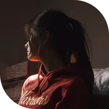 Side profile of a woman with a ponytail wearing a red sweatshirt looking out a window with soft light illuminating her face.