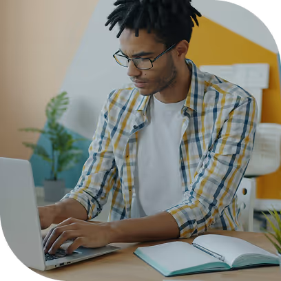 Young man wearing glasses and a plaid shirt typing on a laptop at a desk with an open notebook.