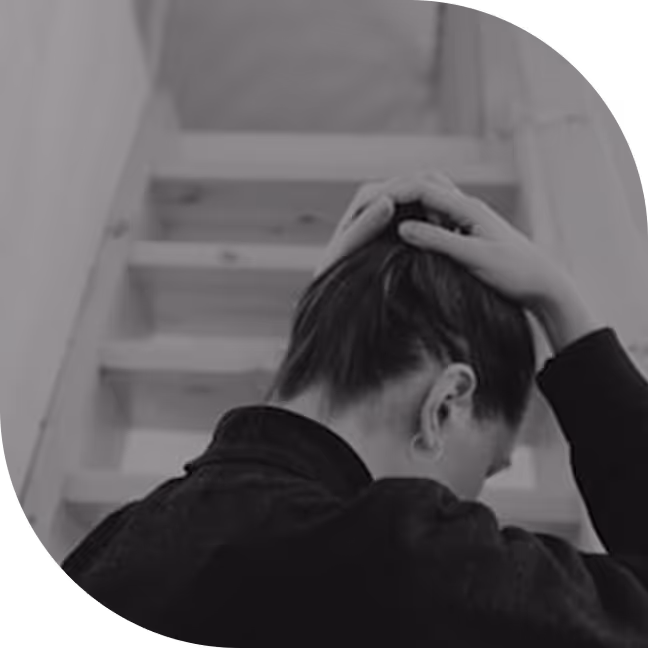 Person with hair tied back holding their head, sitting in front of a staircase, shown from behind.