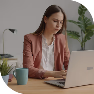 Woman in a brown blazer concentrating while typing on a laptop at a wooden desk with a green mug and potted plant nearby.