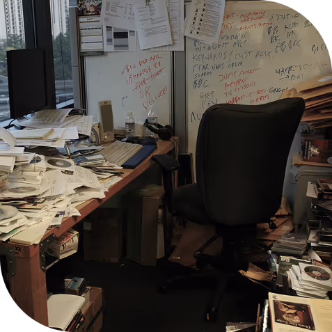Cluttered office space with a black swivel chair, desk piled with papers and CDs, and a whiteboard covered in handwritten notes.