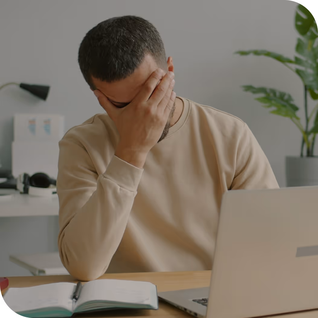 Man in beige sweatshirt sitting at a desk with a laptop and notebook, covering his face with one hand showing frustration.