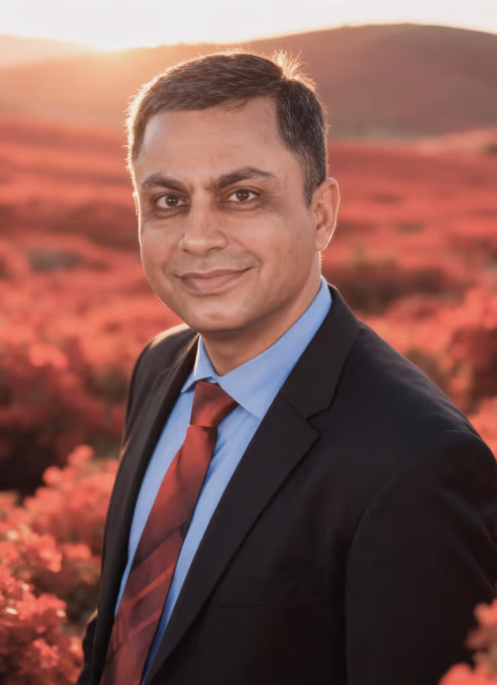 Portrait of a man in a dark suit, blue shirt, and red tie standing outdoors with red flowers and hills in the background at sunset.
