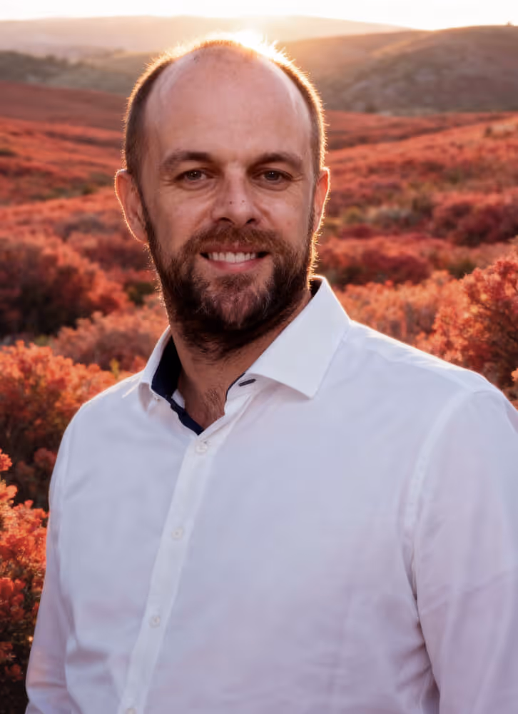 Smiling bald man with a beard wearing a white shirt standing outdoors with hills and red autumn foliage in the background at sunset.