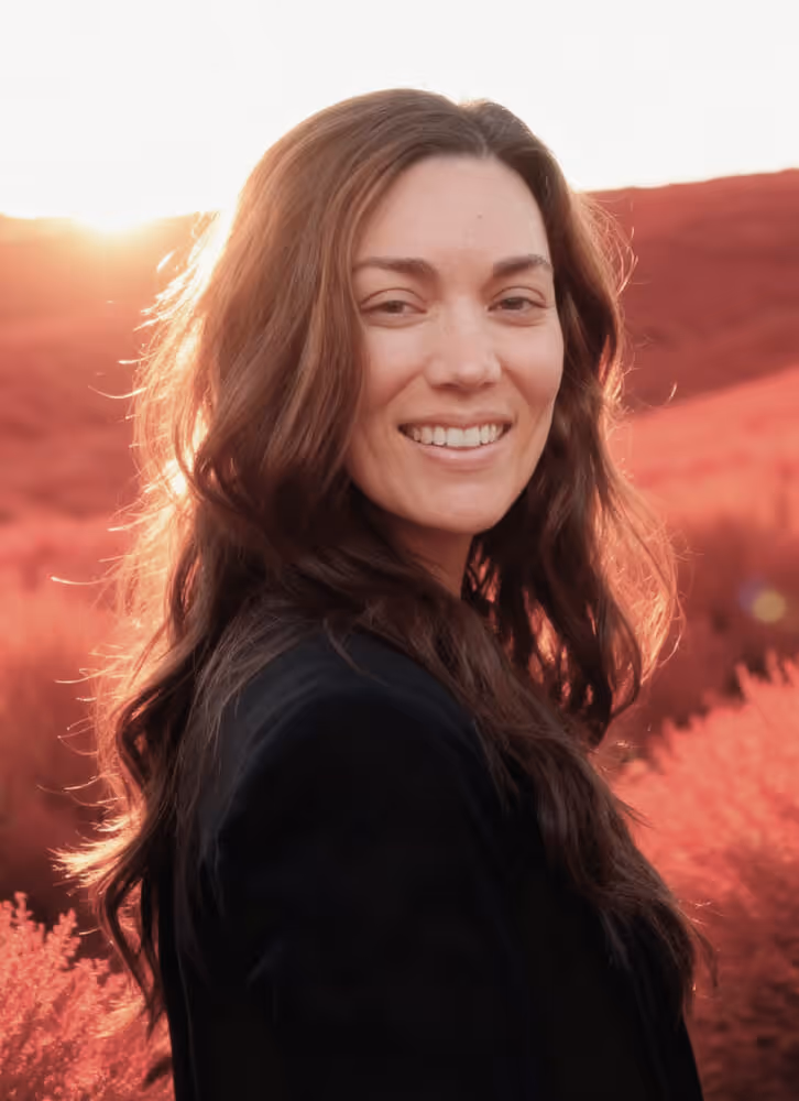 Smiling woman with long wavy brown hair standing outdoors with sunlit red bushes in the background.