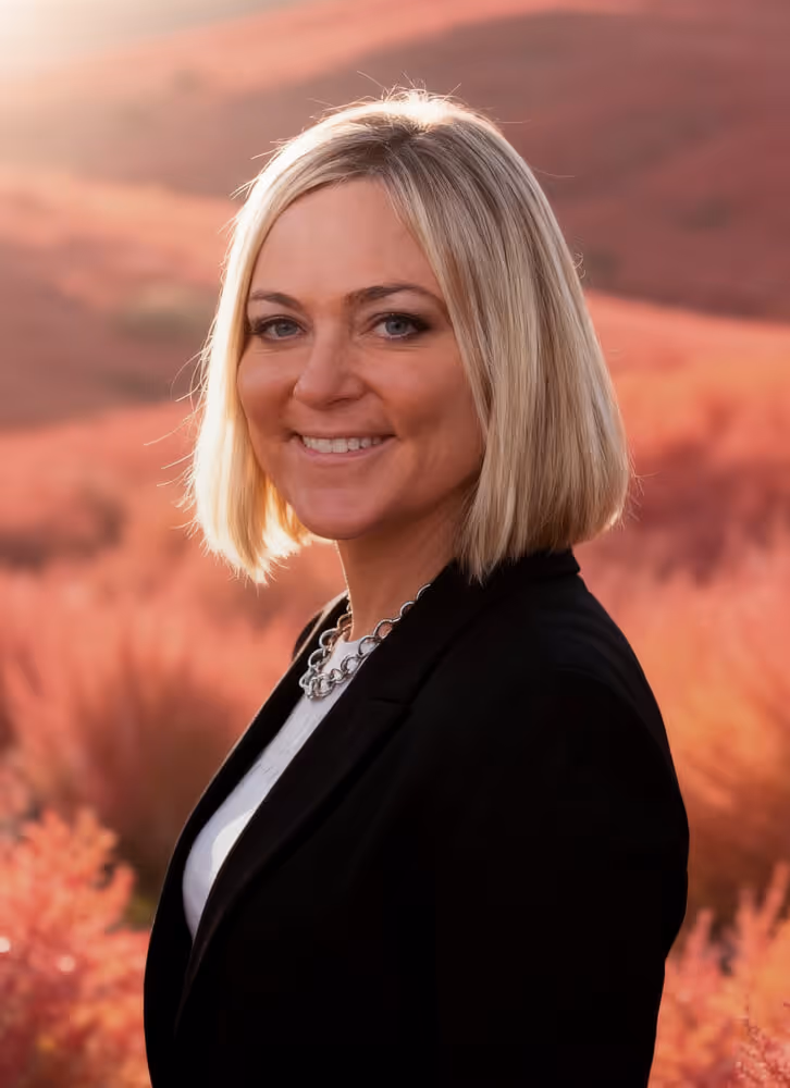 Smiling woman with blonde bob haircut wearing a black blazer and silver necklace, standing outdoors with a blurred reddish background.
