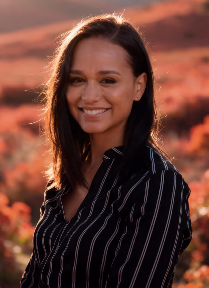 Smiling woman with dark hair wearing a black and white striped shirt in an outdoor setting with warm, blurred foliage background.