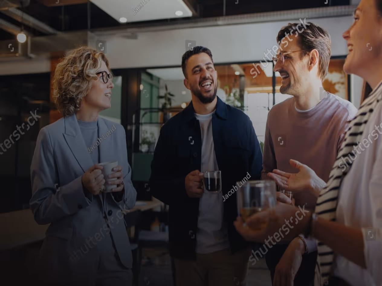 Group of four colleagues standing and chatting while holding coffee mugs in a modern office.