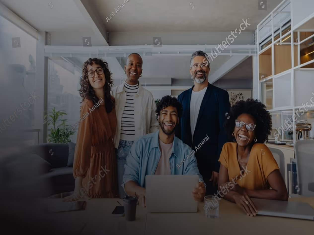 Five diverse colleagues smiling and posing together at a modern office desk with laptops.