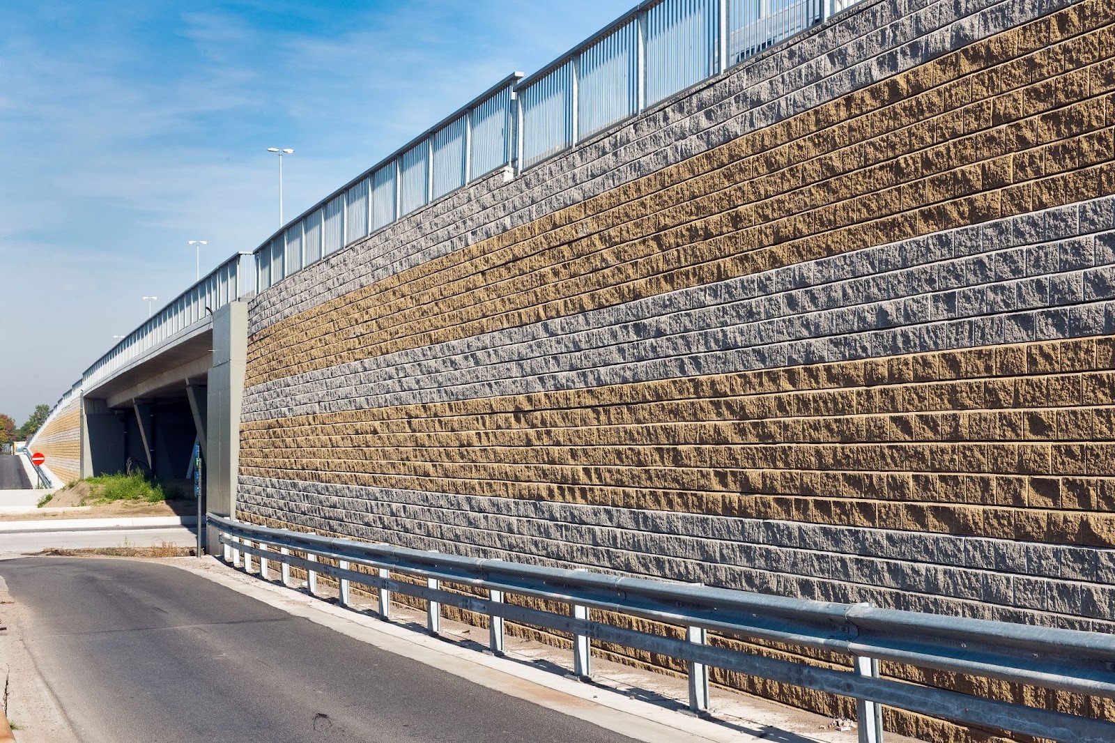 Allan Block retaining wall of interlocking concrete blocks in grey and yellow bands, supporting a highway beside a road and overpass