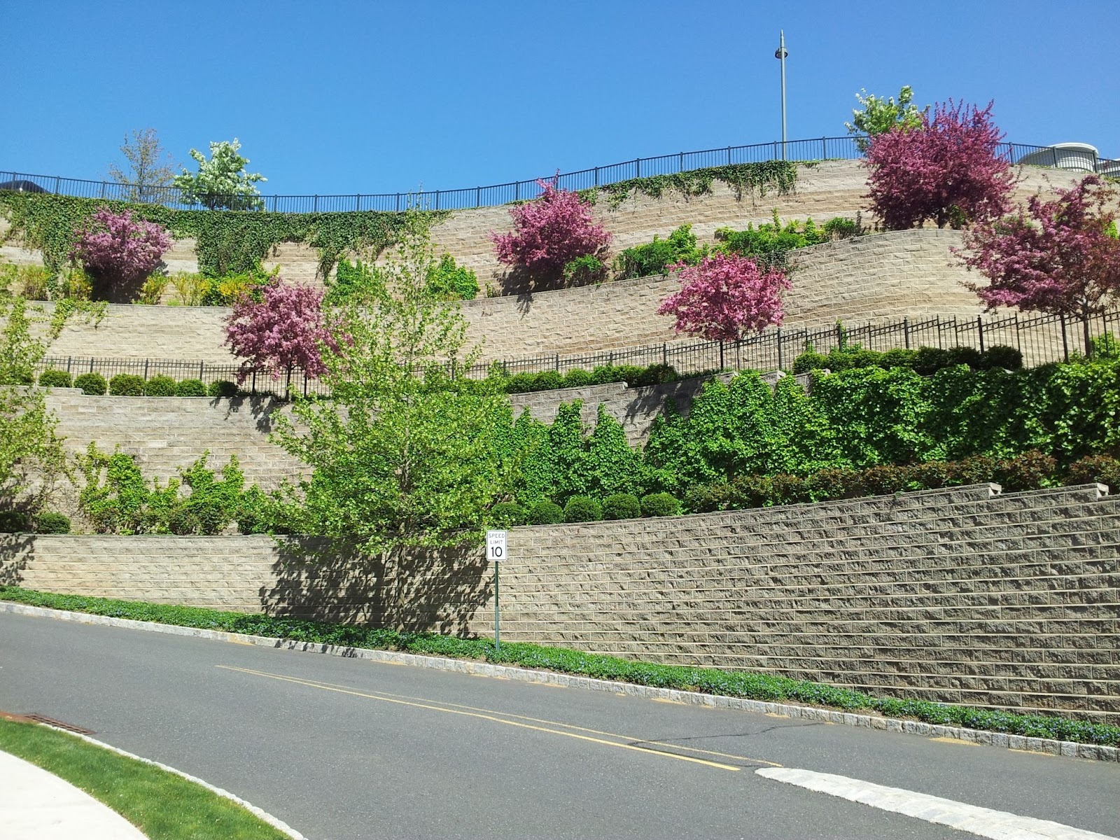 Terraced Allan Block retaining walls with split-face blocks supporting planted slopes and flowering trees along a roadside