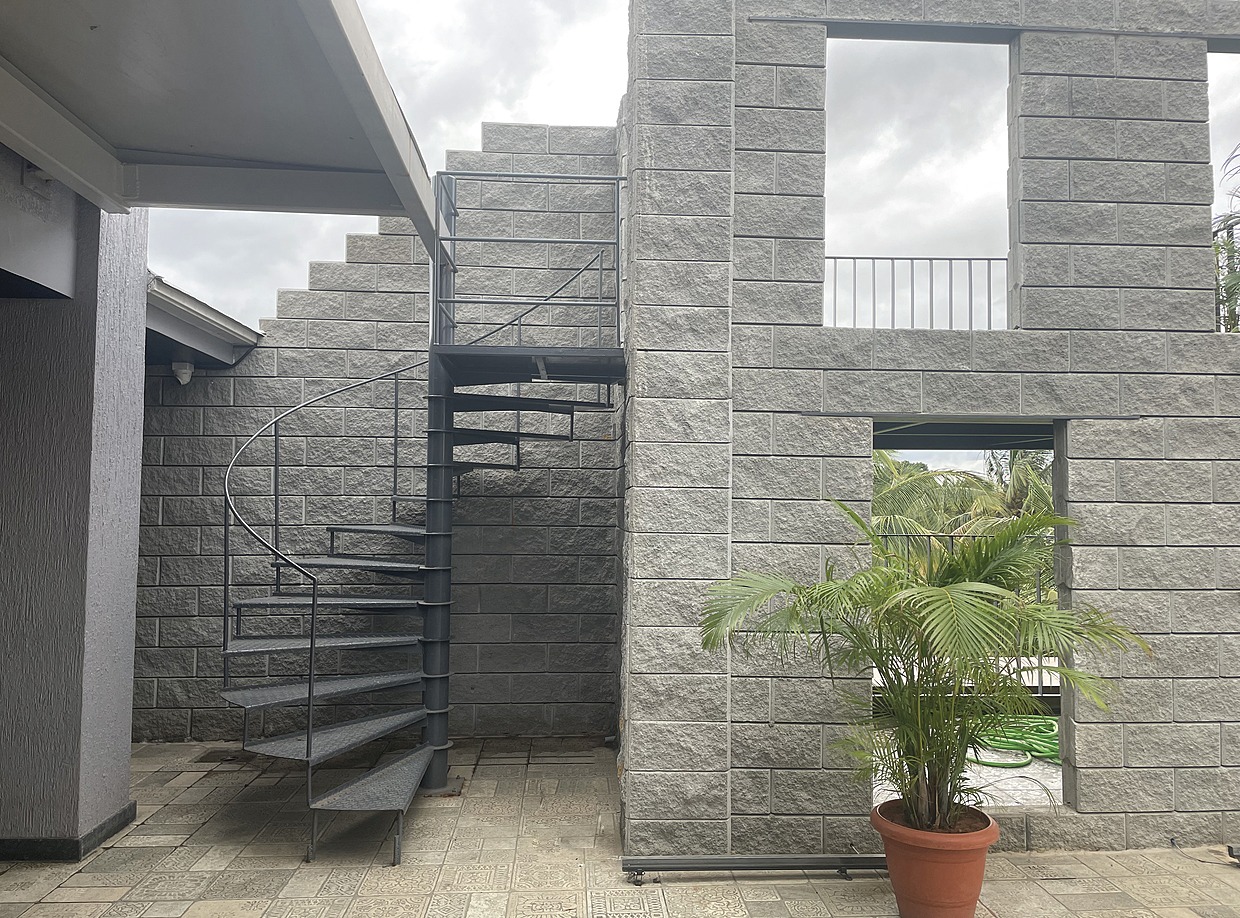 Grey split-face concrete block walls with window openings, beside a spiral metal staircase and a potted palm on paved ground
