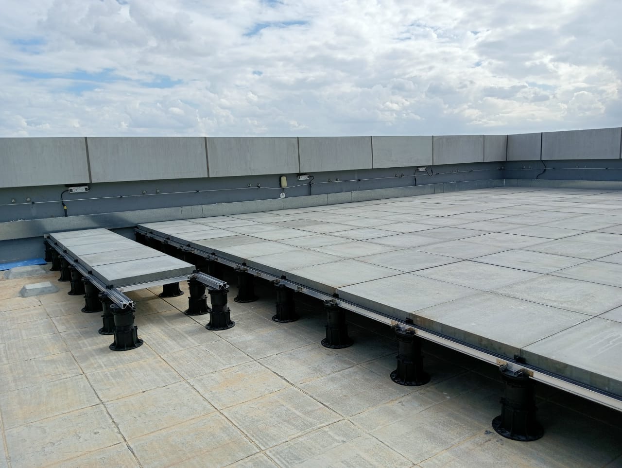 Rooftop terrace of large grey paving tiles raised on adjustable pedestal supports, with some tiles lifted to show the pedestals