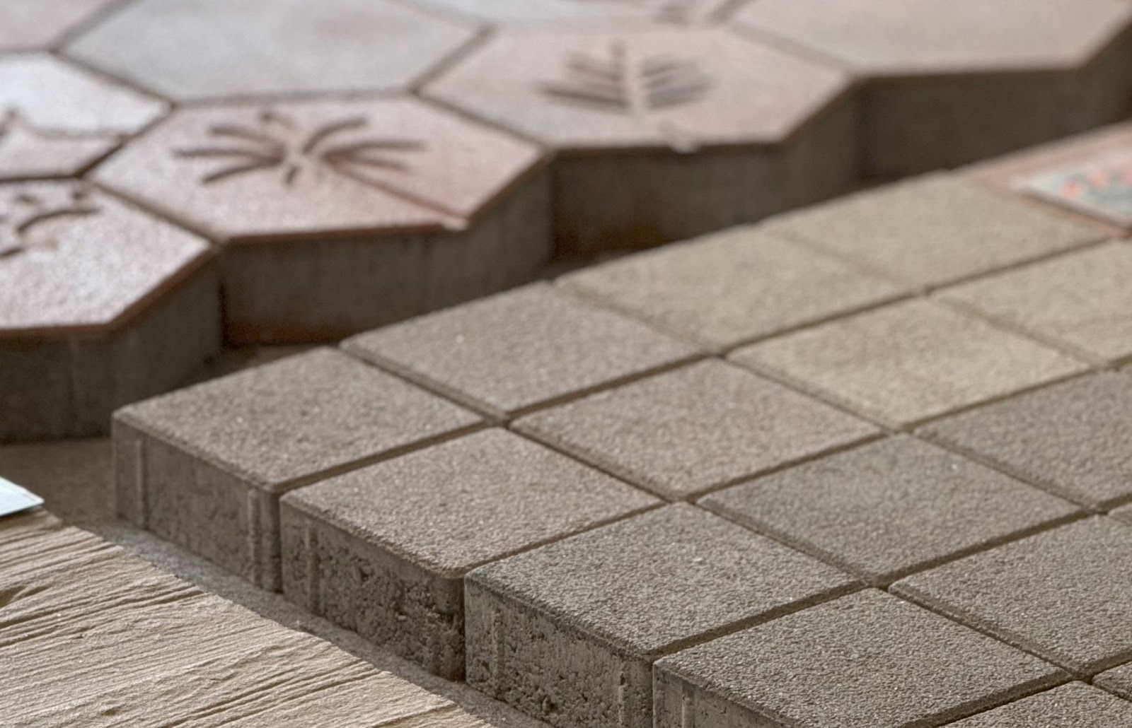 Close-up of square grey concrete pavers next to hexagonal terracotta pavers with leaf motifs, showing their depth and edges