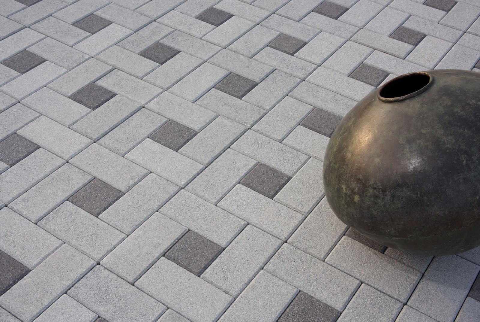 Light grey rectangular concrete pavers in a basket-weave pattern with charcoal square inserts, with a dark metal pot placed on top