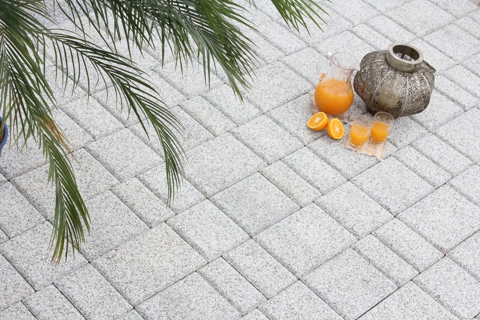 Speckled grey rectangular concrete pavers in a herringbone pattern with a brass pot, oranges and juice glasses under palm fronds