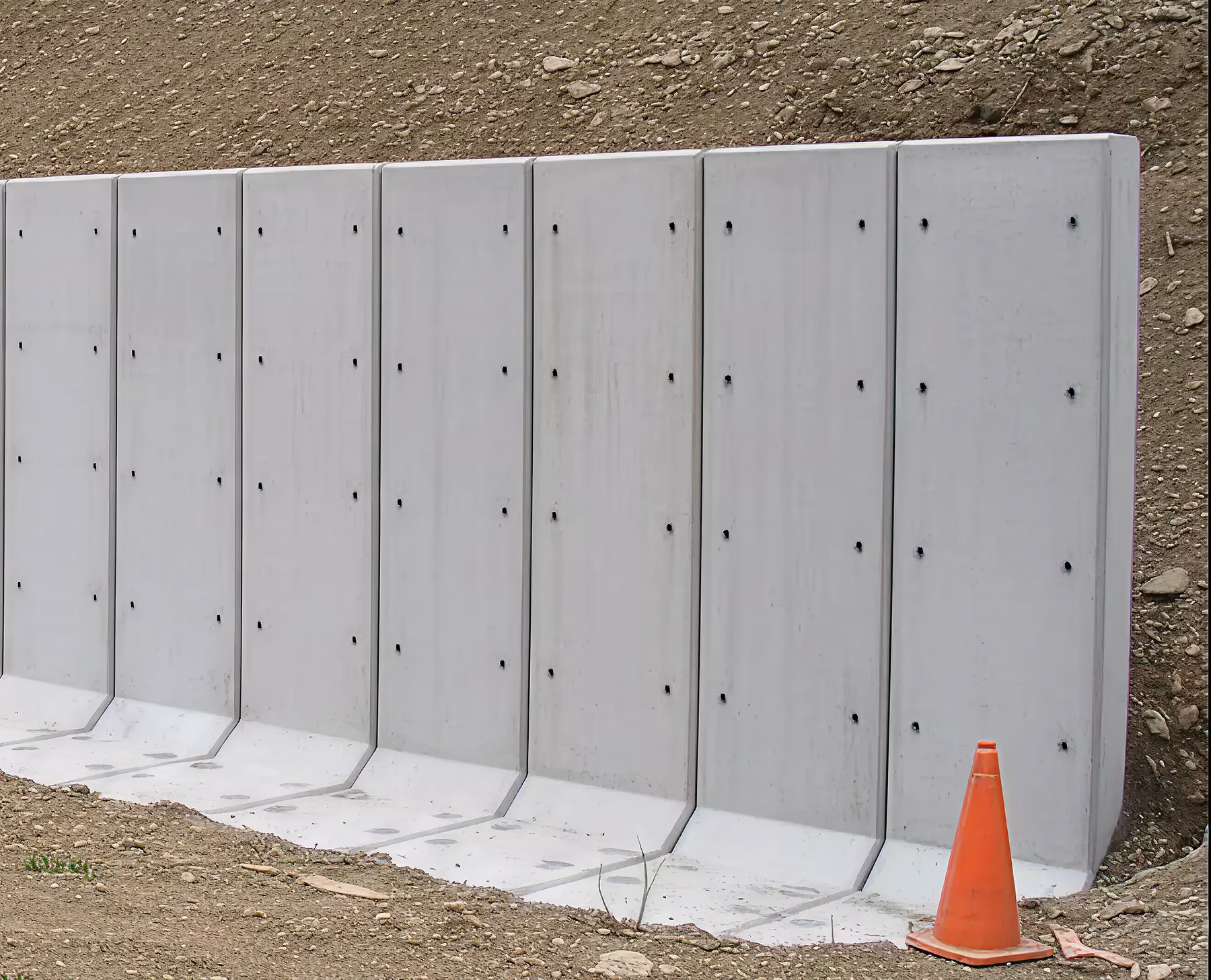 Row of grey precast concrete retaining wall panels standing on a prepared gravel base, with an orange traffic cone at one end