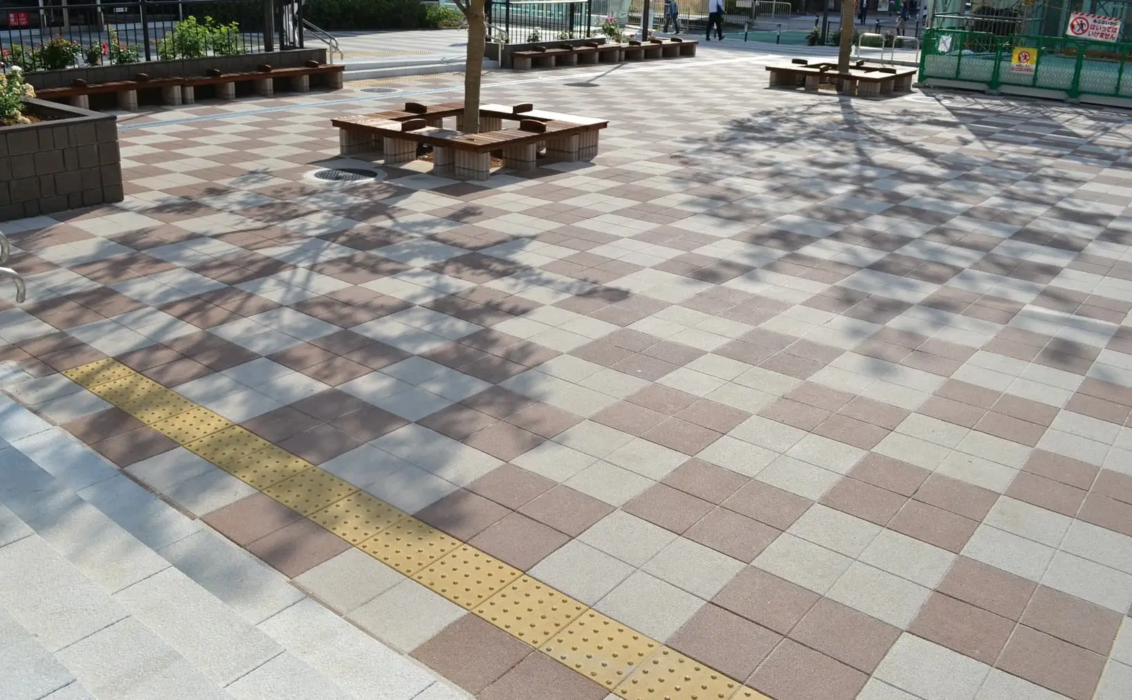 Yellow tactile warning paving tiles with raised dots crossing a beige and red checkerboard plaza with tree seating