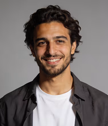 Young man with curly dark hair and light beard smiling, wearing a white shirt and dark jacket against a gray background.