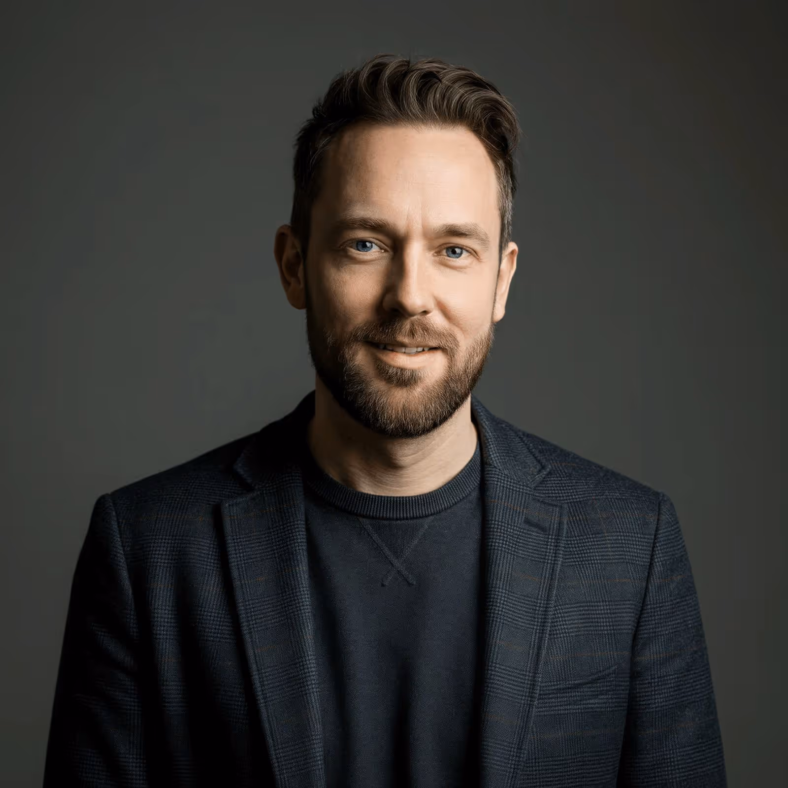 Smiling man with light beard and blue eyes wearing a dark plaid blazer over a black shirt against a dark background.