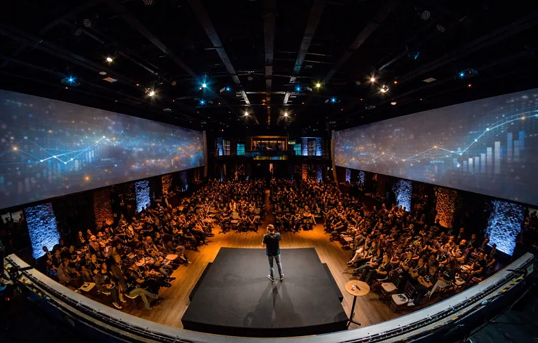 Man standing on a stage addressing a large audience in a dark auditorium with digital graphs projected on wide screens.