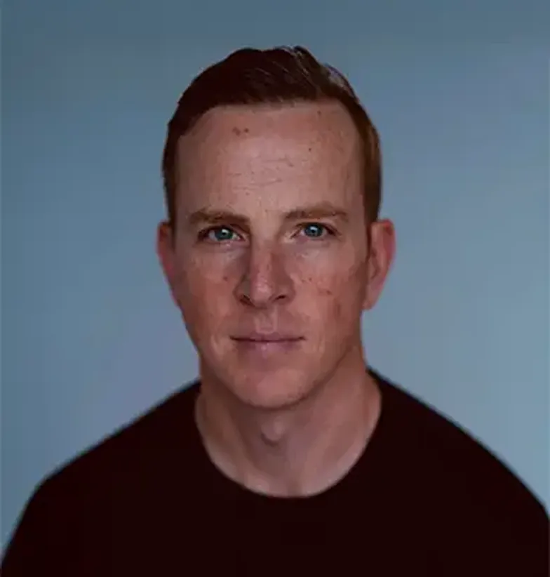 Close-up portrait of a man with short brown hair and blue eyes wearing a black shirt against a plain blue background.