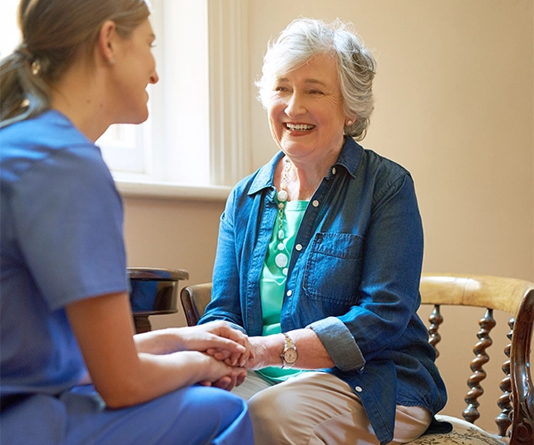 Smiling elderly woman holding hands with a nurse dressed in blue scrubs in a warmly lit room.