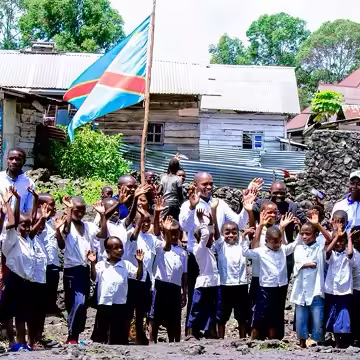 Group of children and adults standing outdoors, some waving, with a large flag and rustic buildings in the background.
