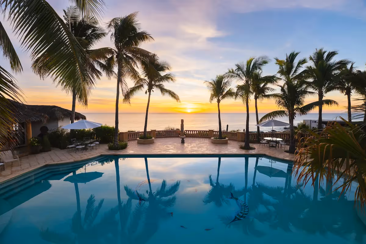 Vista del atardecer sobre la piscina infinita rodeada de palmeras en el restaurante del hotel La Valise Los Cabos con vista al océano.