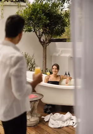 A couple enjoying a relaxing moment at La Valise Mexico City hotel, featuring a woman in a freestanding bathtub under an indoor tree while a man brings her a drink.