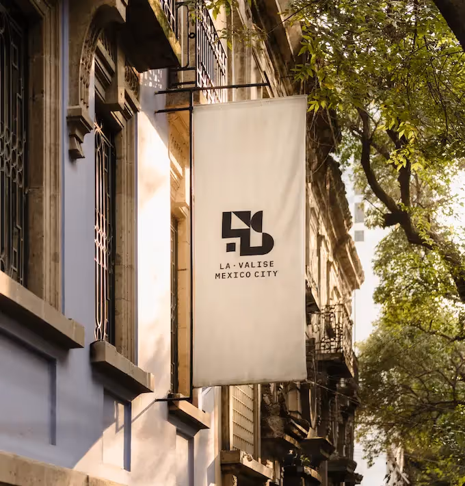 White exterior hanging sign of La Valise Mexico City hotel on a tree-lined street.