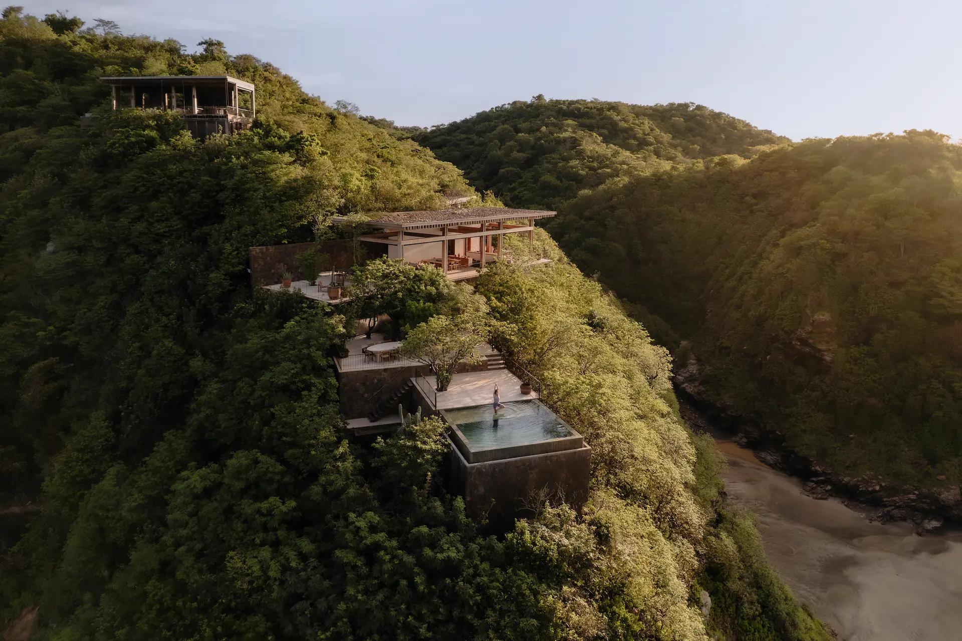 Modern house perched on a lush green hillside with an infinity pool overlooking a river valley at sunset.