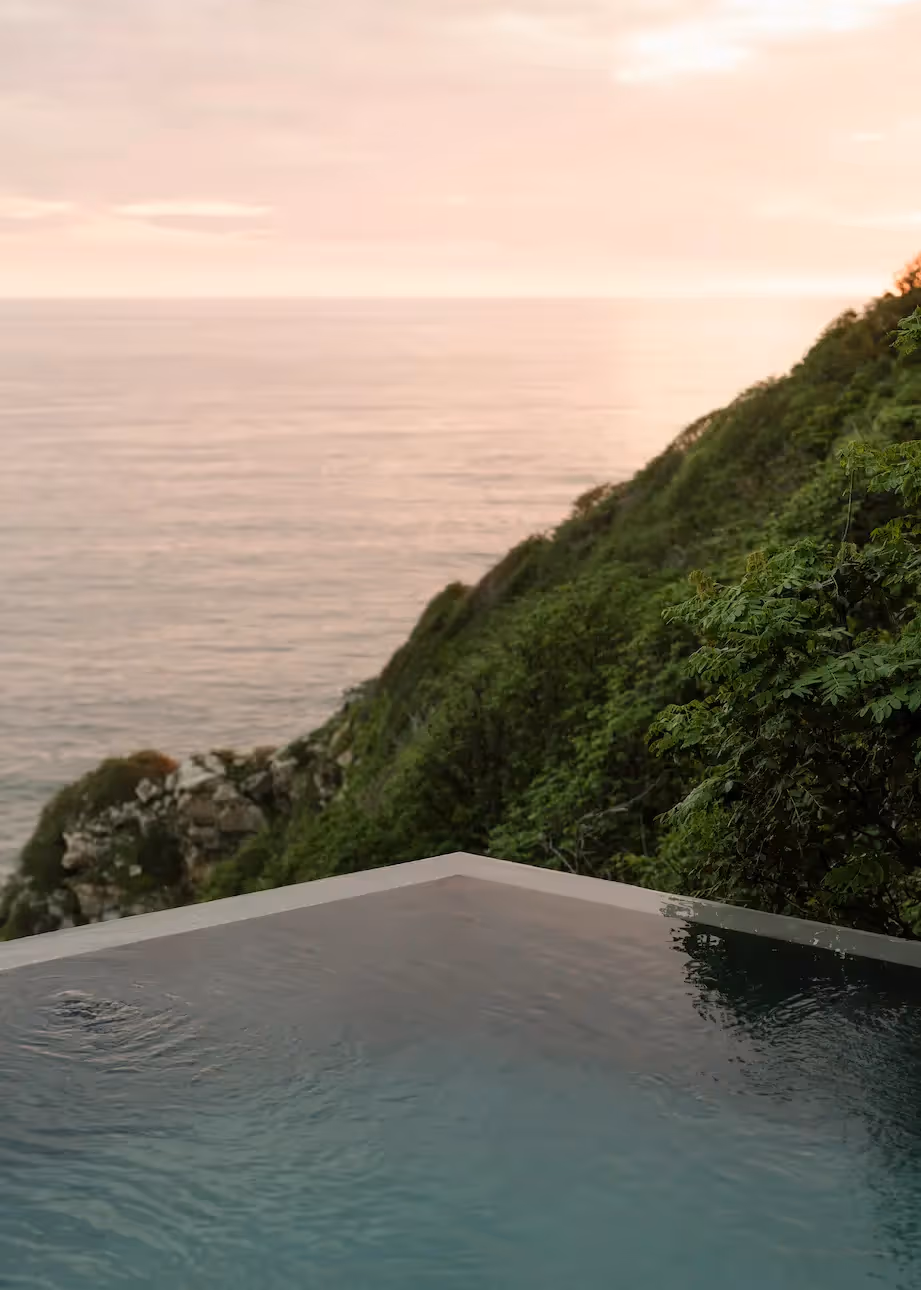 Infinity pool at La Valise Mazunte Hotel overlooking the Pacific Ocean and green hills at twilight.