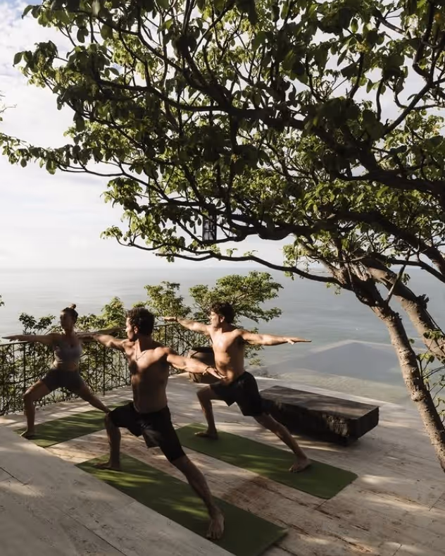Three people practicing yoga on a panoramic terrace at La Valise Mazunte Hotel, overlooking the ocean under the shade of tropical trees.