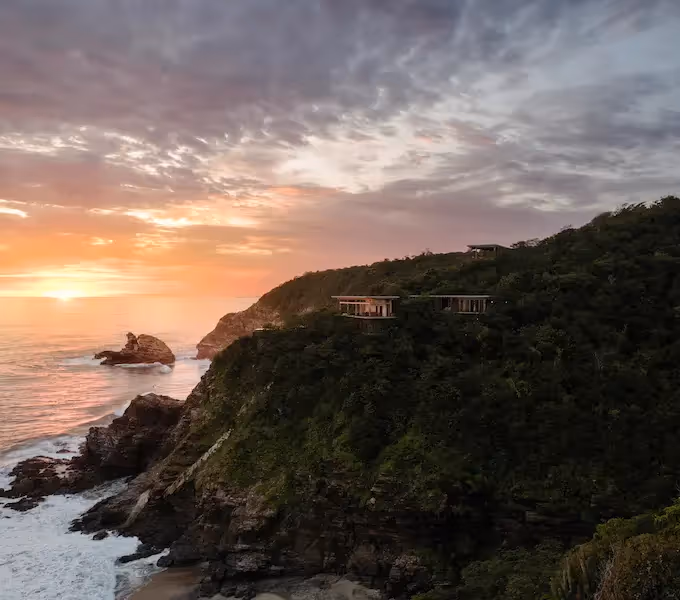 Una vista cautivadora de la puesta de sol del océano, con un acantilado cubierto de vegetación en primer plano, formaciones rocosas y el restaurante del hotel La Valise Mazunte.