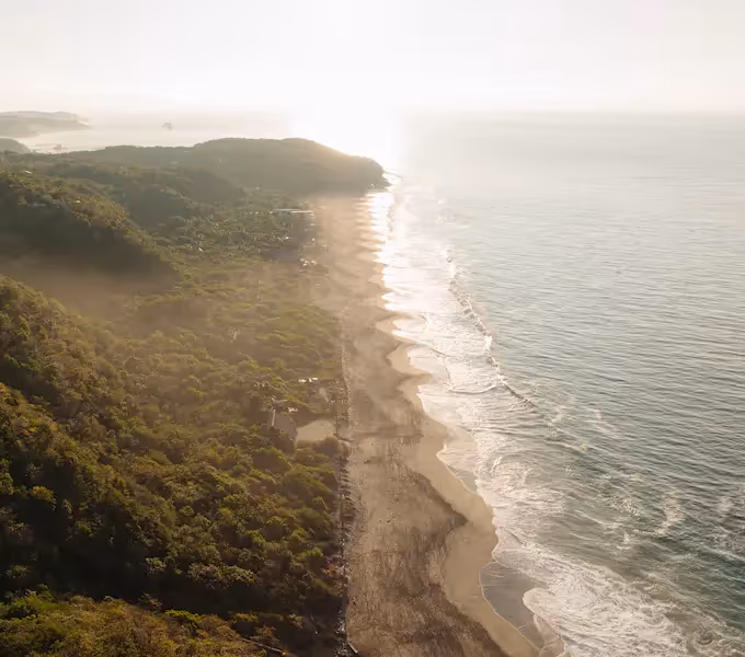 A bird's eye view of a secluded beach at sunrise, with the La Valise Mazunte Hotel restaurant nestled in the cliffside, overlooking the ocean.
