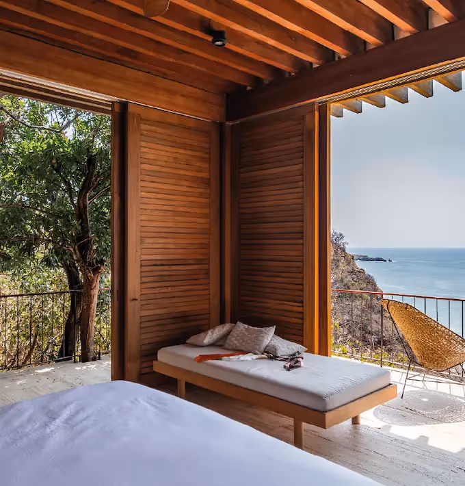 Panoramic view from a suite at La Valise Mazunte Hotel featuring a luxurious bed, a private wooden terrace with rustic furniture, palm trees, and the Caribbean Sea through a large thatched A-frame structure.