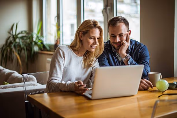 A smiling couple sitting at a wooden table looking at a laptop together in a bright room with large windows.