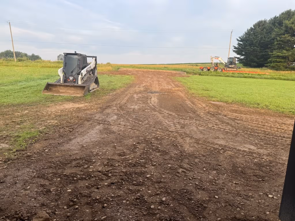 Dirt and gravel driveway under construction with a compact track loader on the left and an excavator in the distance on the right.